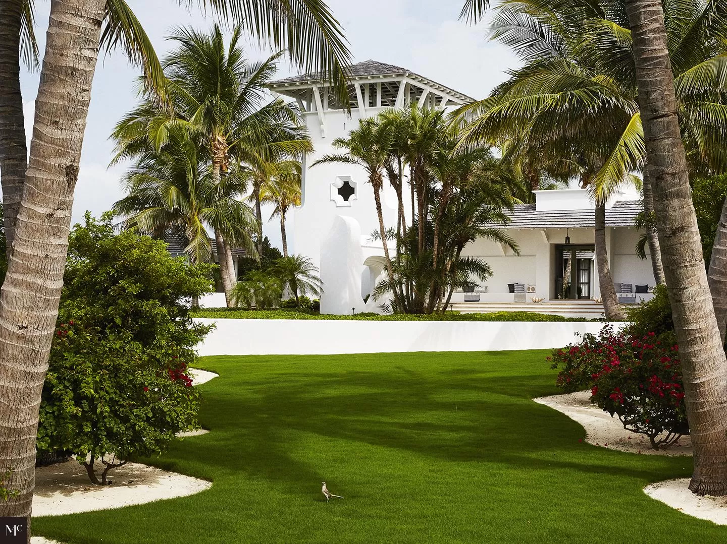 A front facade of an all-white bungalow with large windows, columns flanking each door, a shingled roof, circular chimney, and tropical plants on a green lawn