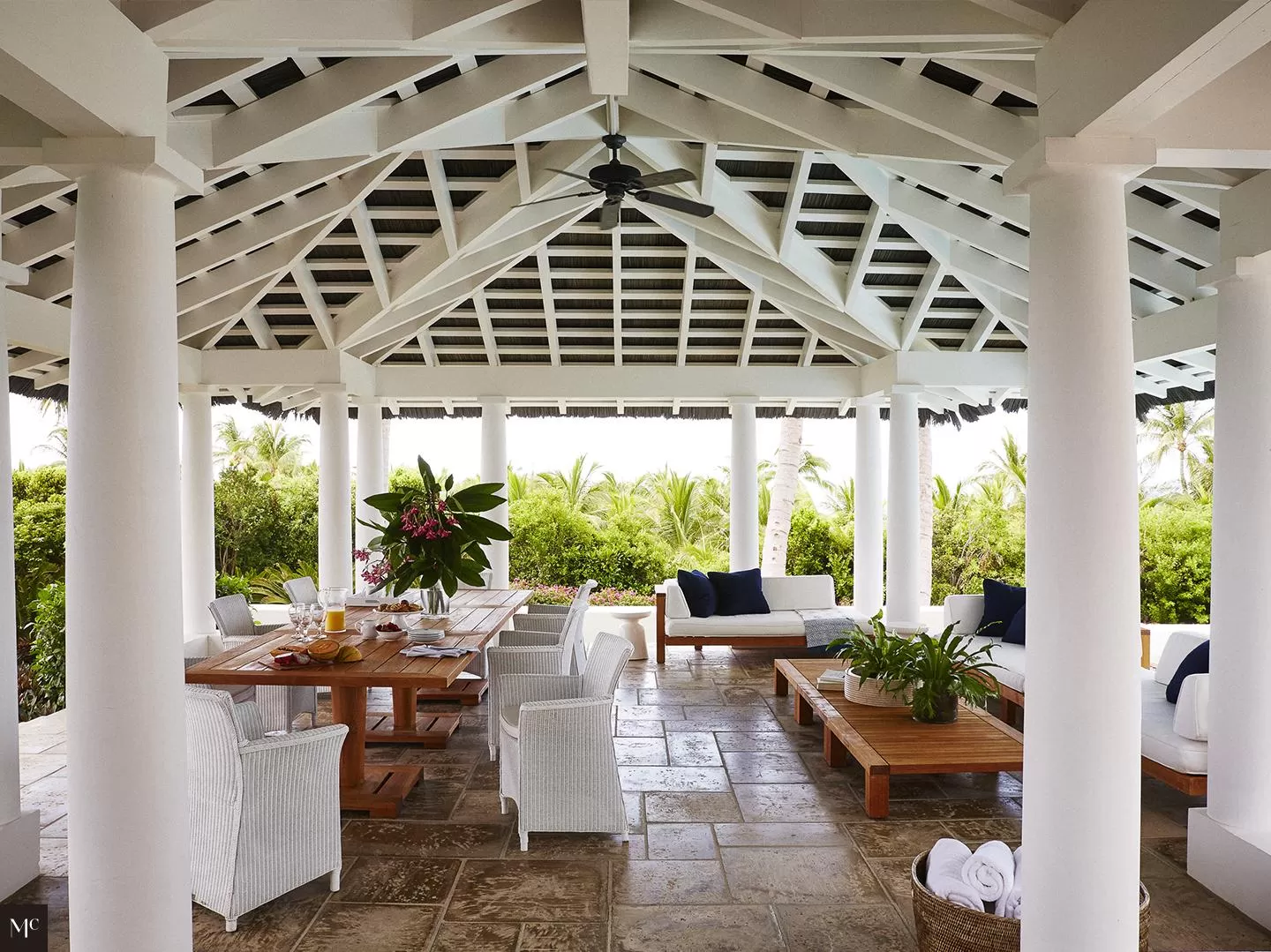 A backyard of an all-white bungalow with columns flanking each door, a shingled roof, circular chimney, overlooking tropical plants on a green lawn