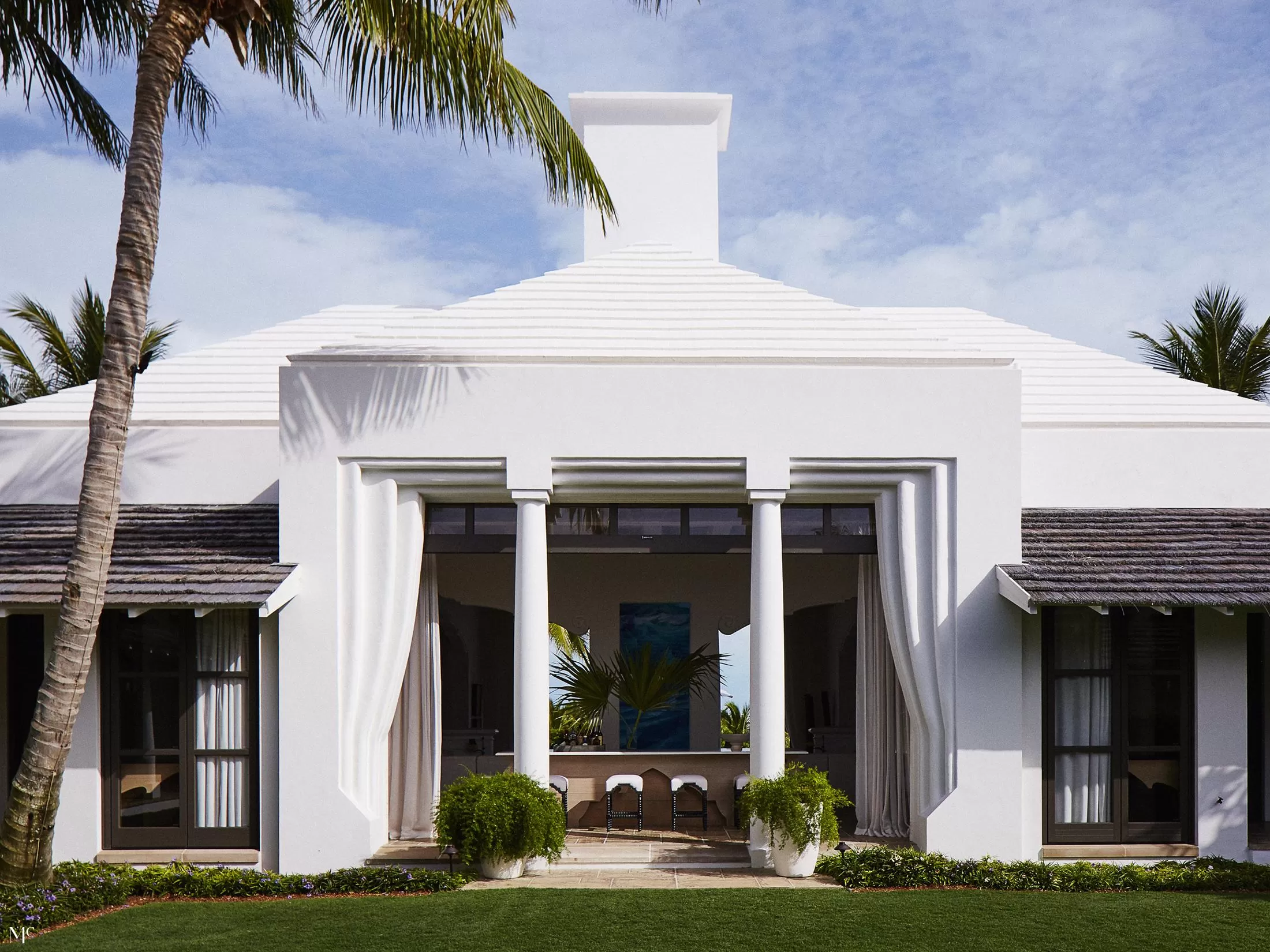 A front facade of an all-white bungalow with large windows, columns flanking each door, a shingled roof, circular chimney, and tropical plants on a green lawn