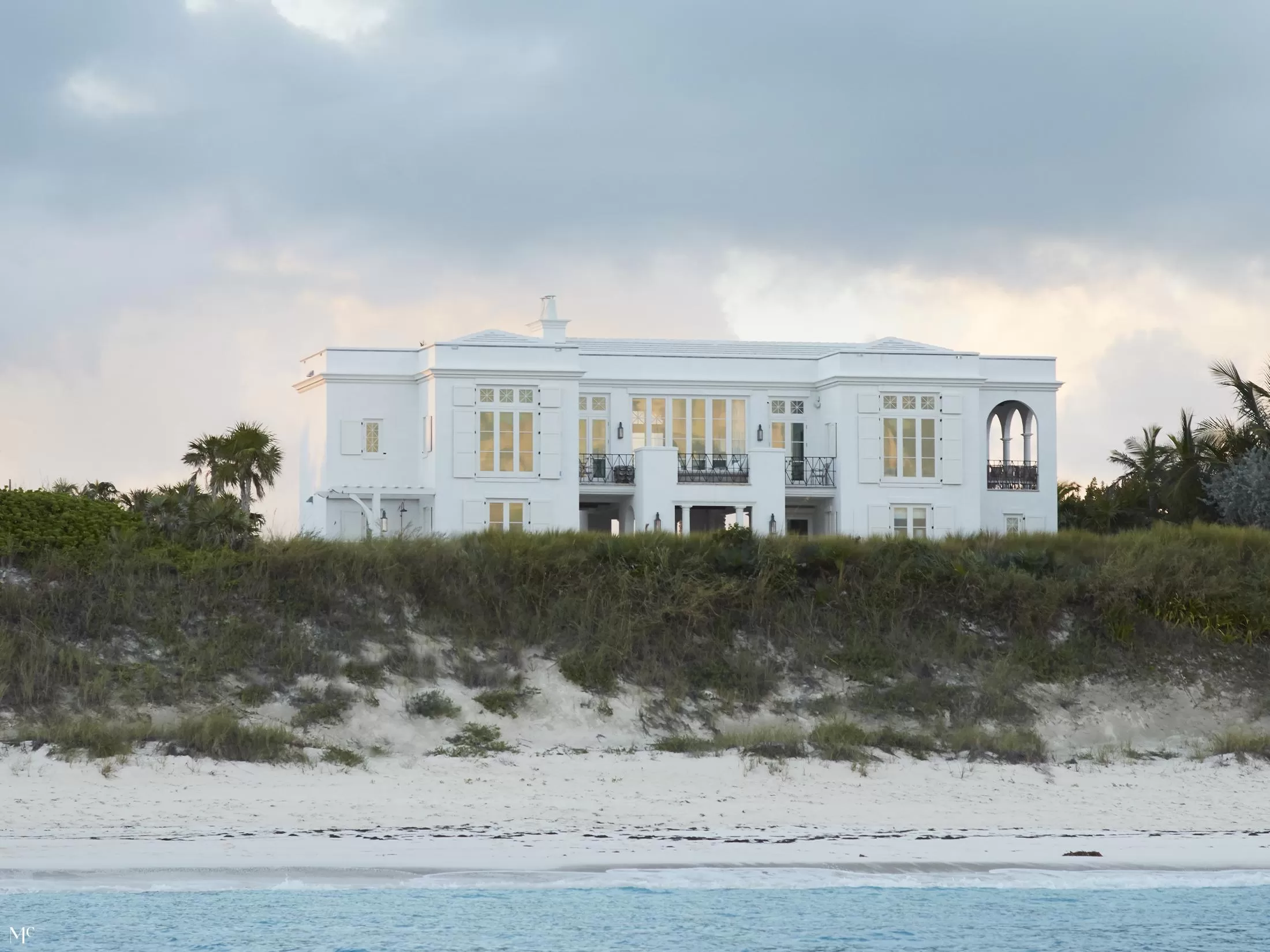 A wide shot of a white beach mansion in with simple design, sand dunes, and the ocean in the background