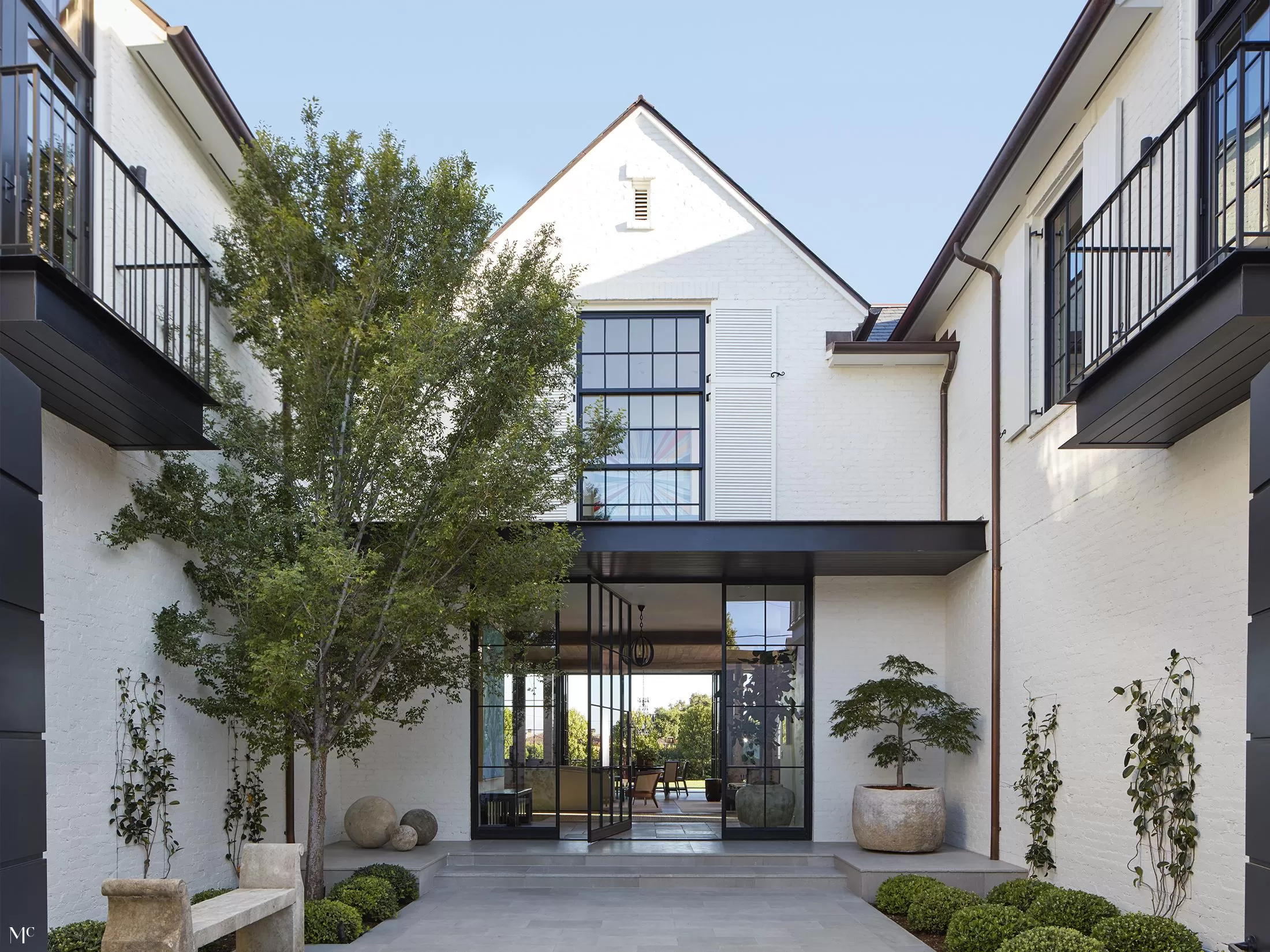 An architectural front view of a modern farmhouse courtyard with white and black buildings, large glass doors, oversized trees, and neutral tones