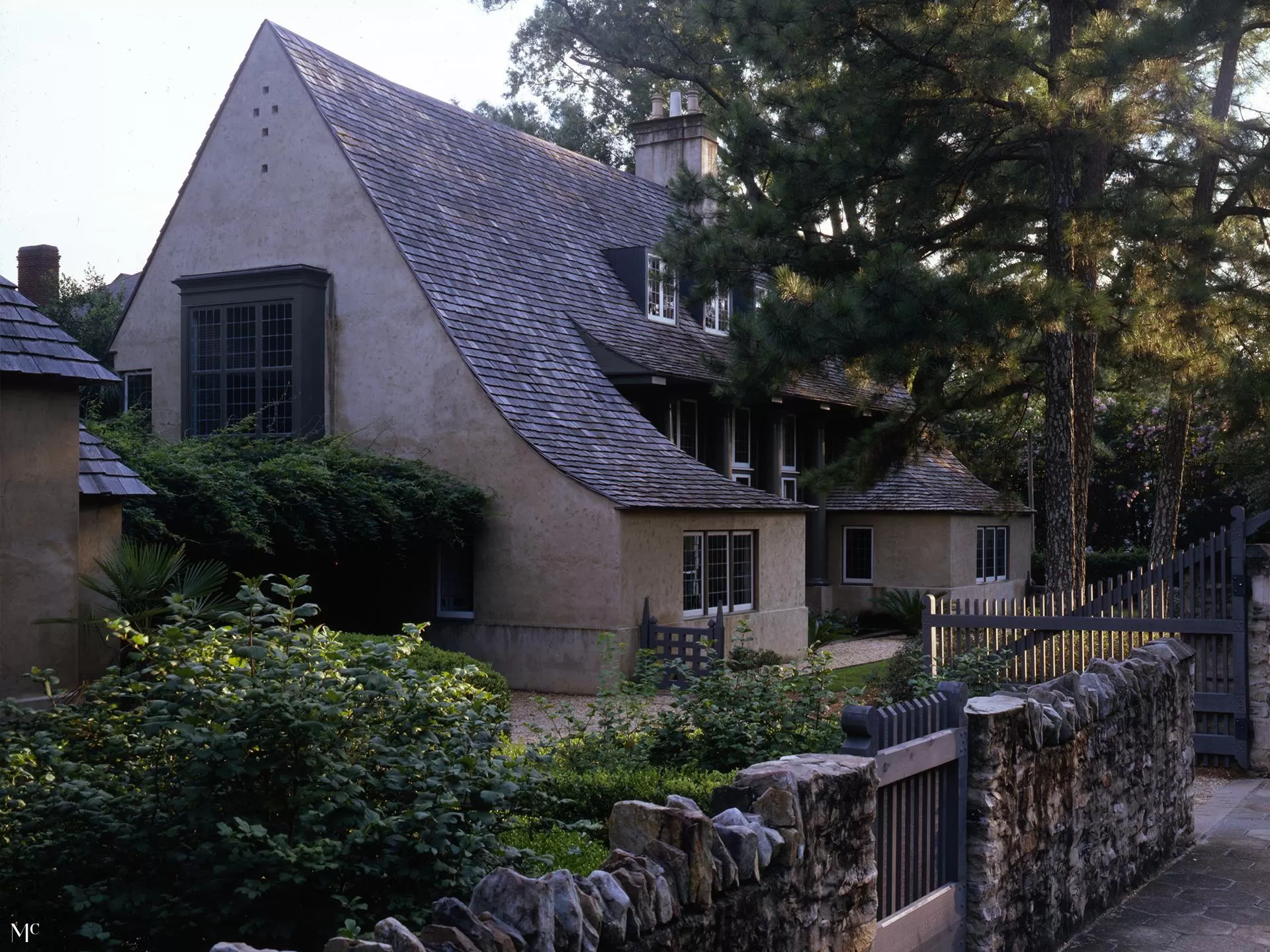 front and side view of an old, rustic cottage with a gray shingle roof surrounded by stone walls, an ivy-covered garden fence, and a black wooden gate leading to a courtyard