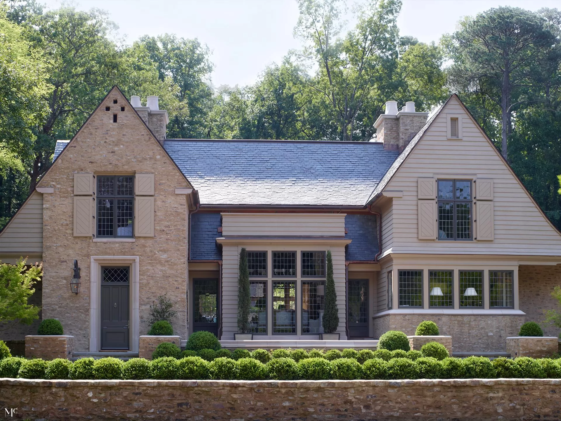 front exterior view of a traditional stone and tan brick home in a pristine, harmoniously designed style, with a blue roof, modern farmhouse aesthetic, large windows, and dark grey shutters, surrounded by lush green trees