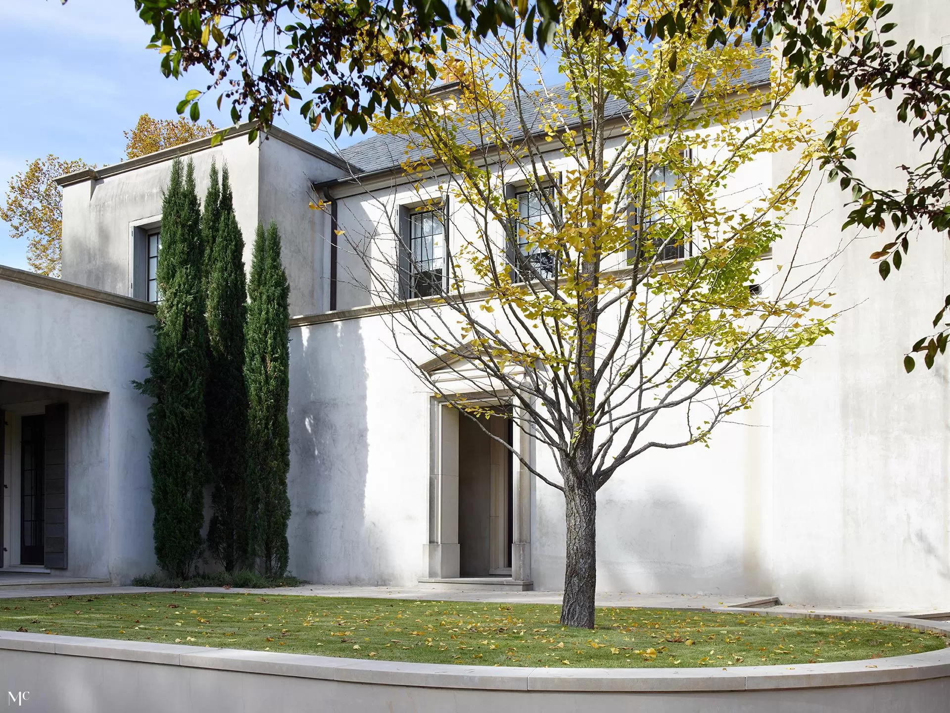 Exterior wall and courtyard, featuring two tall cypress trees on either side of an entrance door to three small rooms with large windows, all in white stucco walls