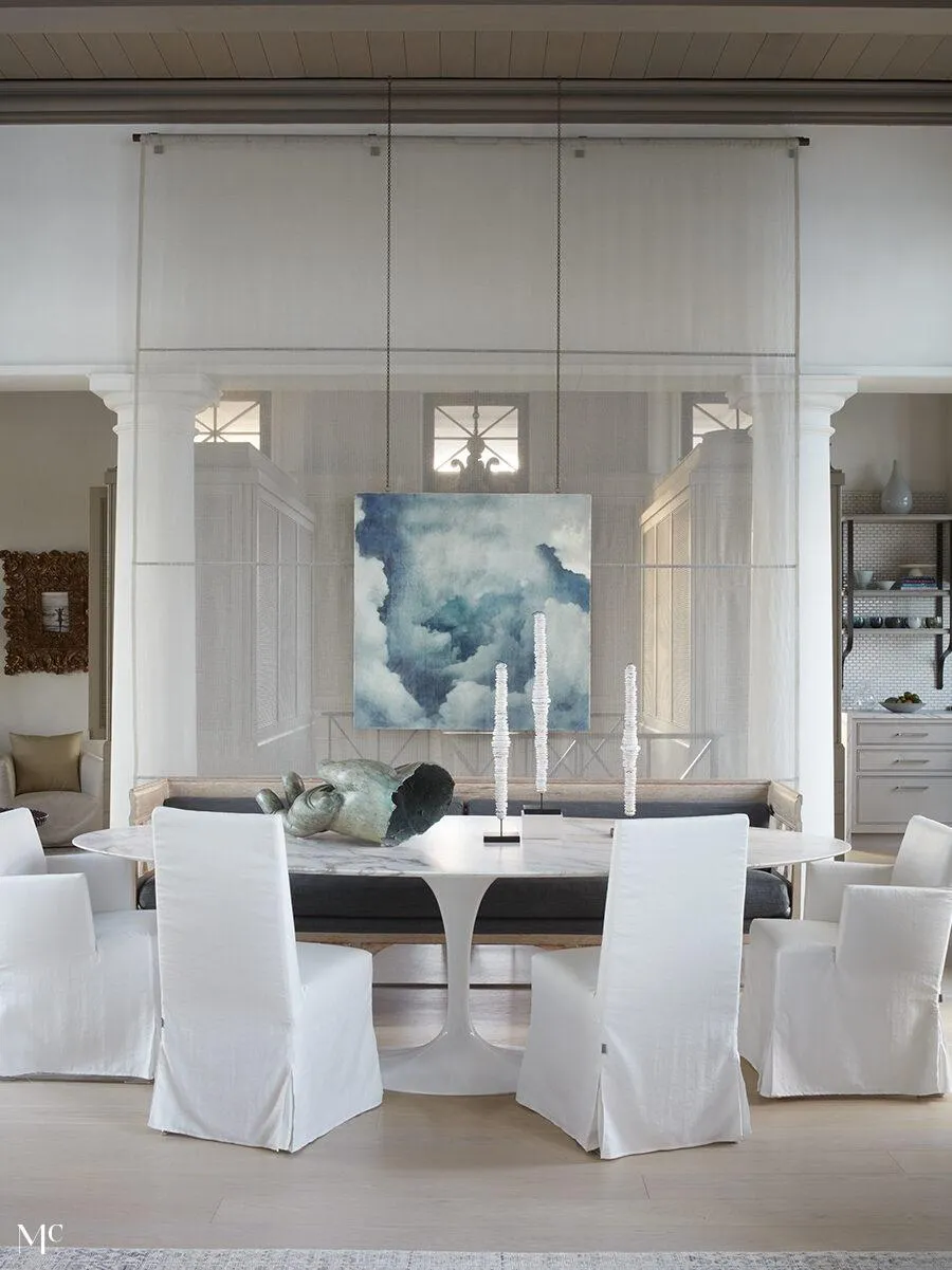 A modern coastal dining room with white chairs, large artwork on the wall above the table, hanging sheer curtains, and natural light, showcasing a neutral color palette and organic textures.