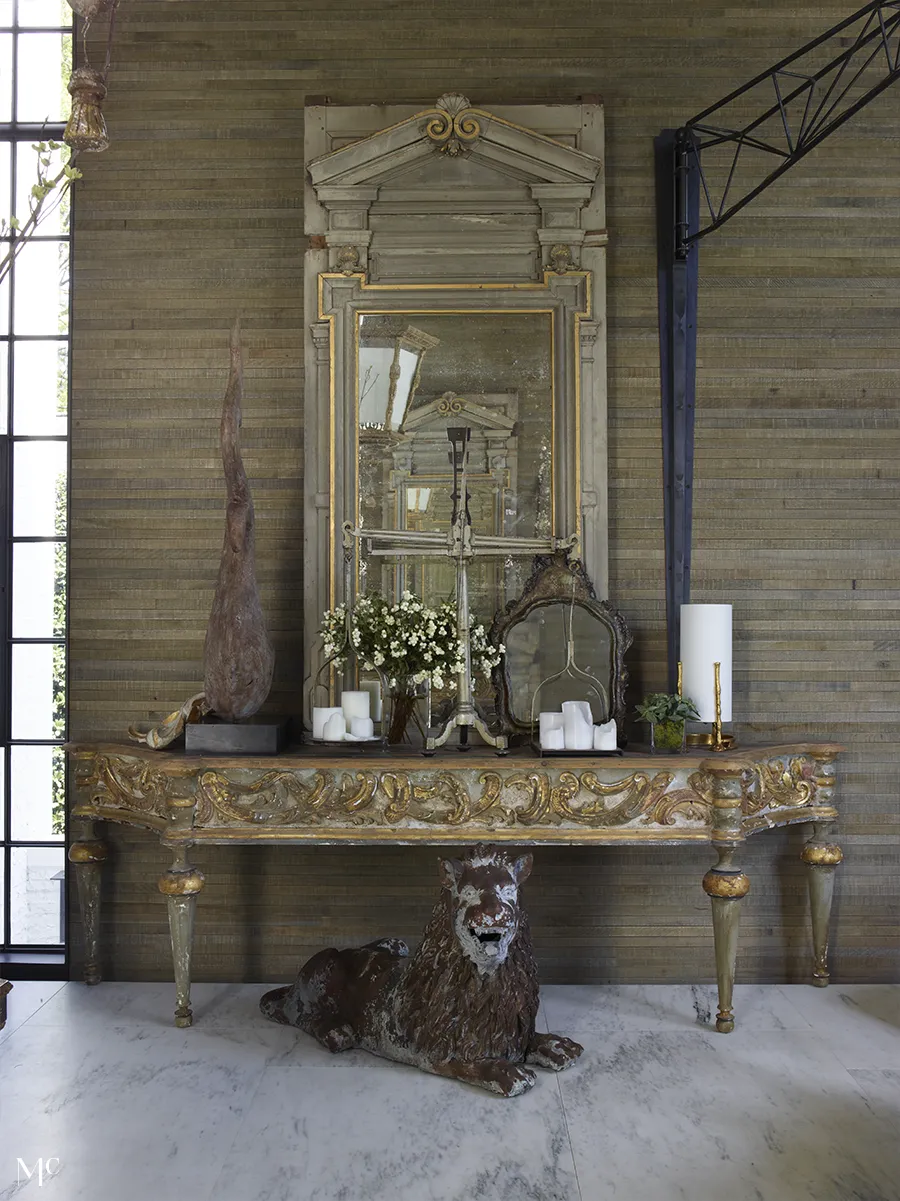 An industrial entryway features a carved console with brass legs, vintage mirrors, bronze sculptures, flowers, and candles, blending rustic charm with luxury.