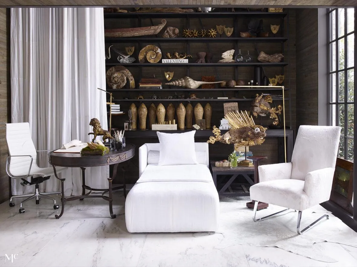 A modern home office with natural light features a white chaise lounge, marble floors, dark wood paneling, industrial shelves filled with artifacts, and a blend of luxury and rustic design, complemented by sculptural accents and comfortable seating.