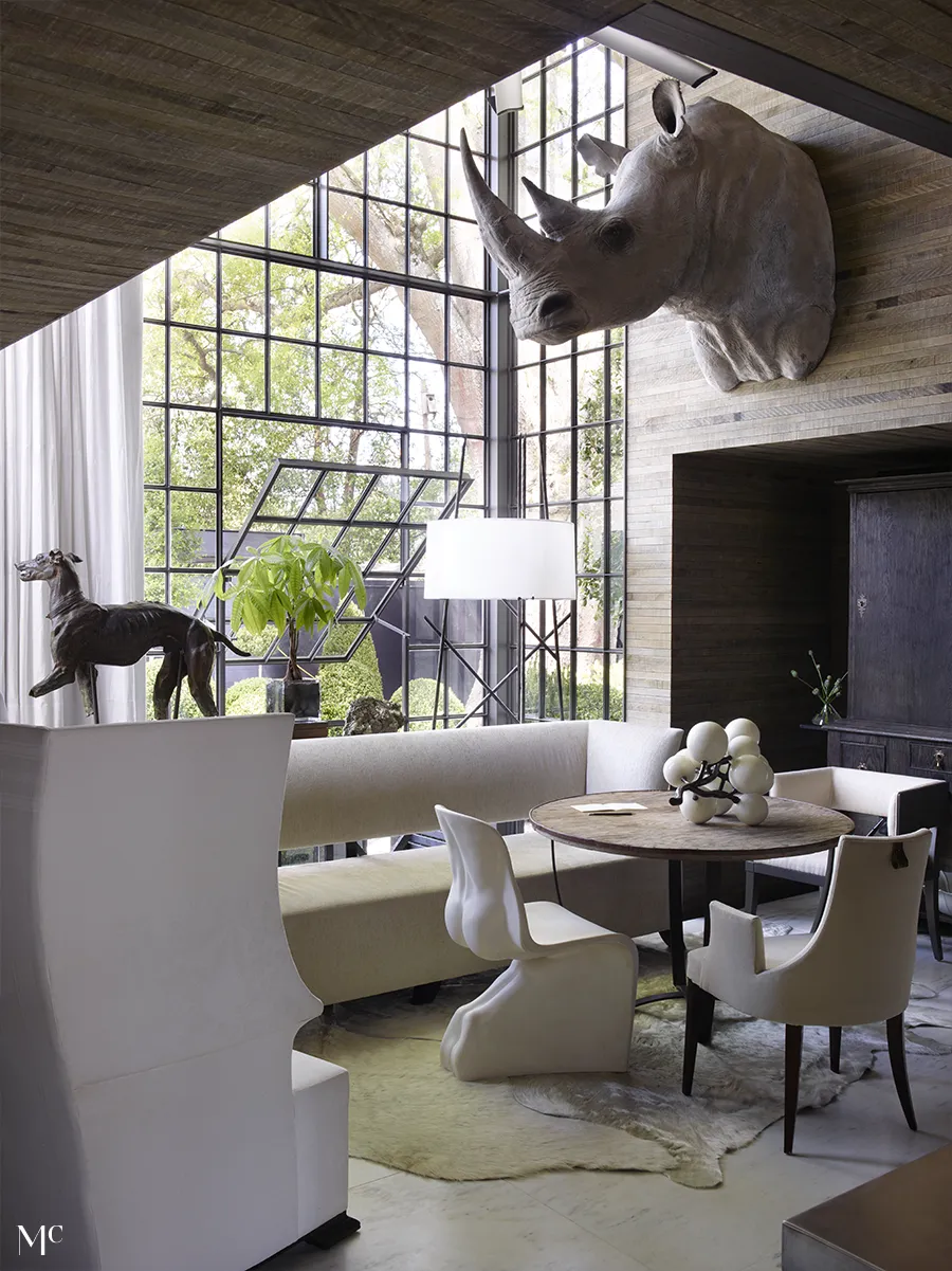 A modern living space with floor-to-ceiling black steel windows, wooden furniture, and sculptures that cast natural light and shadows, framed by a view of trees and nature outside.