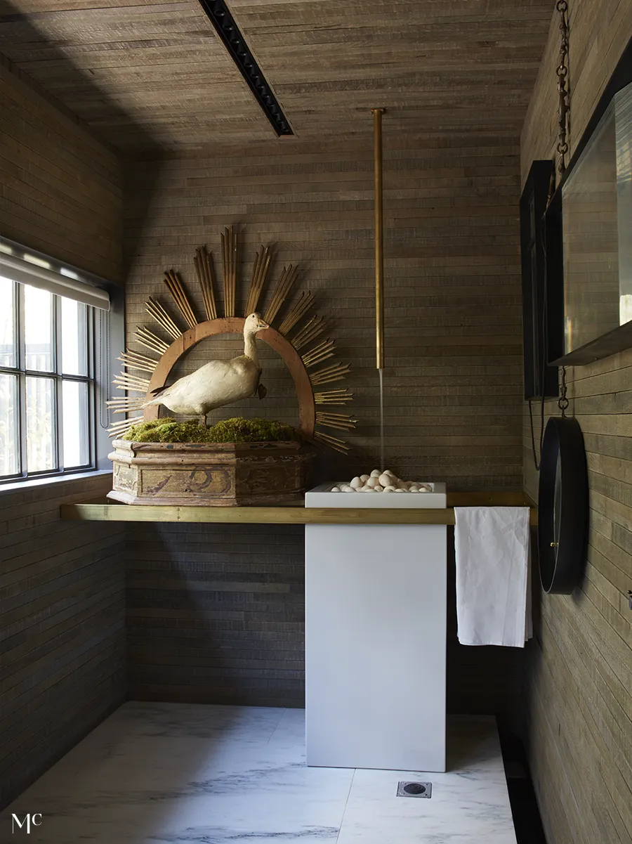 A modern bathroom with marble floors, wood walls, brass accents, and a moss-covered goose statue, blending nature with sleek contemporary design.