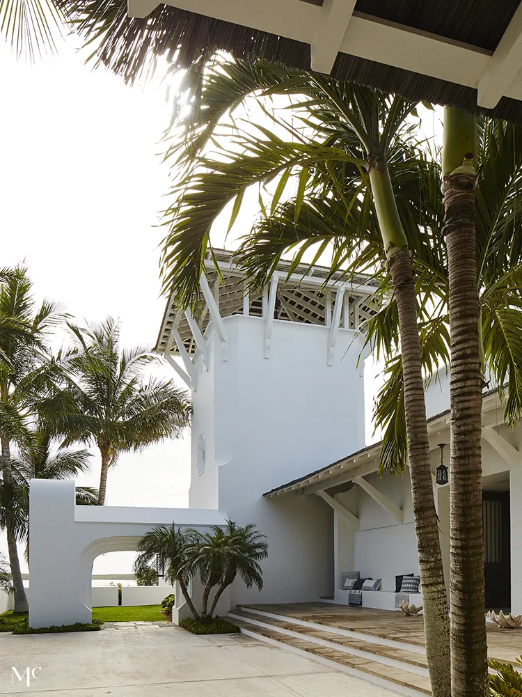 A side facade of an all-white bungalow with large windows, columns flanking each door, a shingled roof, circular chimney, and tropical plants on a green lawn