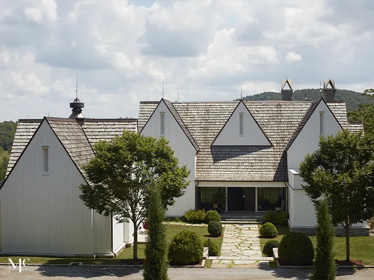 A large, white New England-style mansion with a gable roof and tall windows sits on a grassy hill overlooking a valley