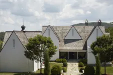 A large, white New England-style mansion with a gable roof and tall windows sits on a grassy hill overlooking a valley