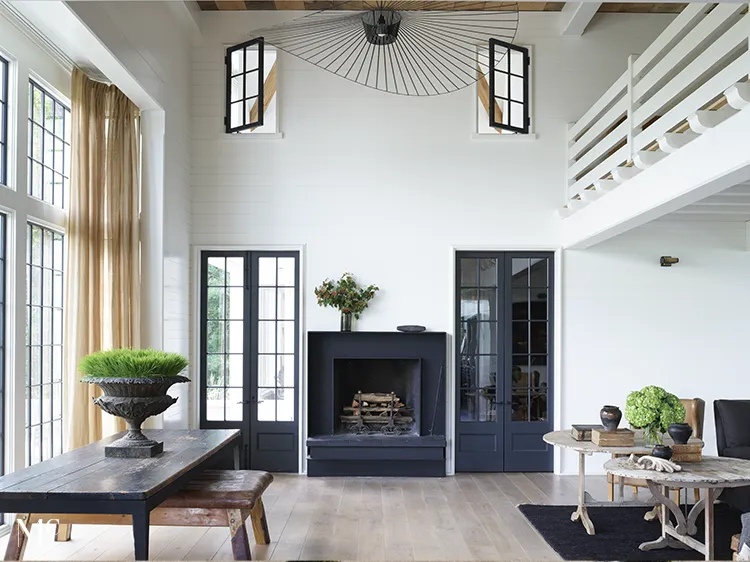 A farmhouse living room with black steel double doors, white oak plank floors, dark windows, full-height black French cabinetry surrounding the fireplace, ceiling beams, brass light fixtures, a large dining table, high-back chairs, and green plants in planters