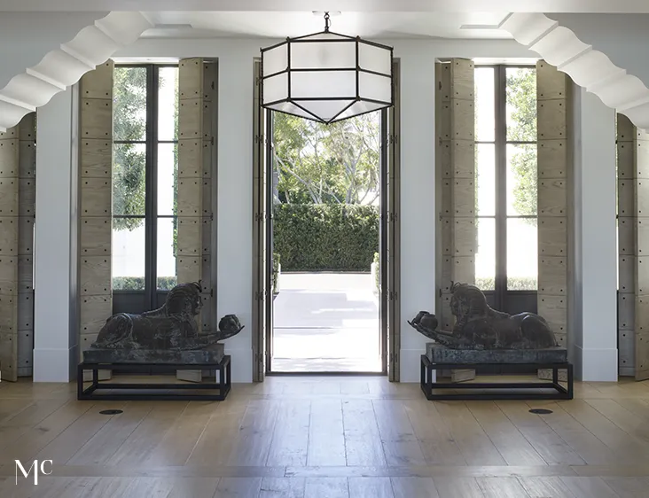 large hallway with double doors leading to a patio, large black metal sculptures on each side of the door, light wood floors, tall windows, and white walls