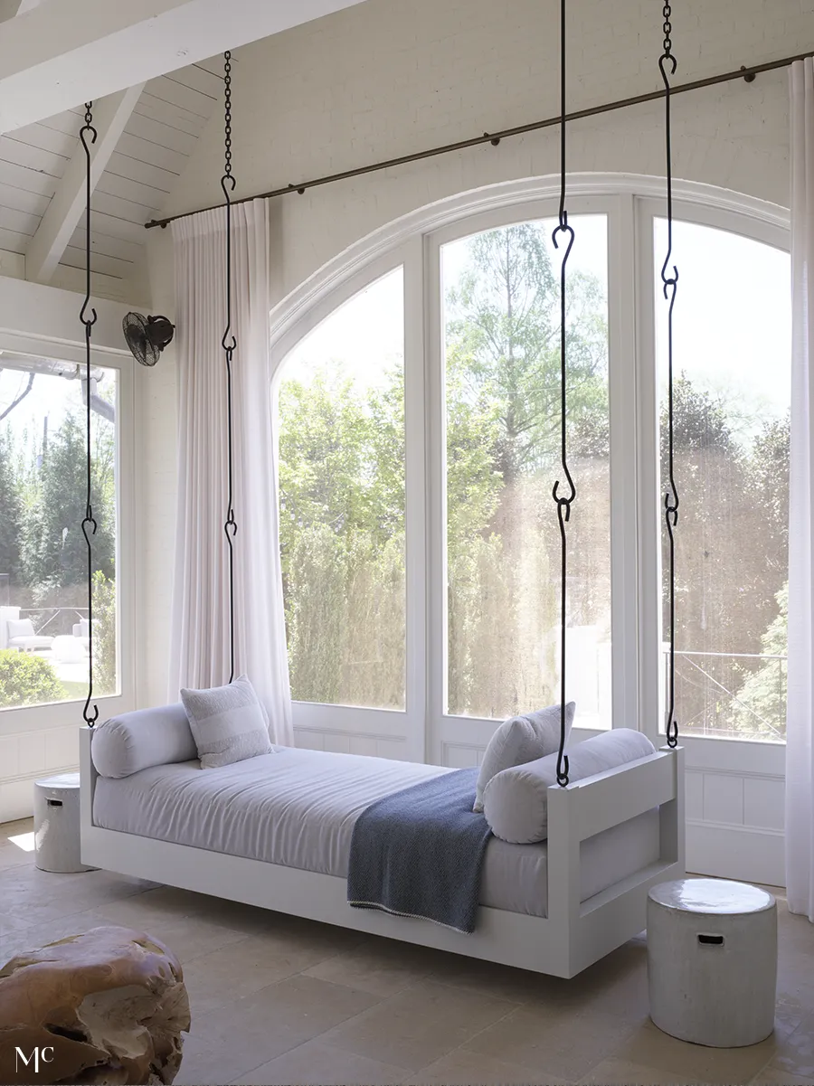 An elegant white swivel secretary desk with a hanging bed, black chains, and gray pillows in front of large windows