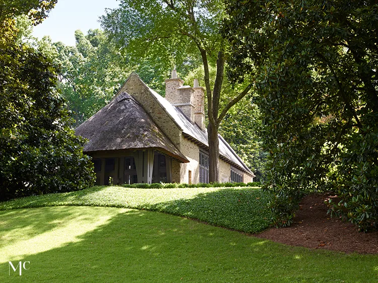 A traditional, two-story country club building with stone walls and a shingle roof stands in the center of an elegant driveway surrounded by trees and steps leading to it