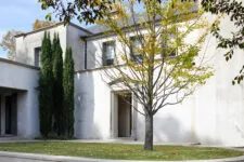 Exterior wall and courtyard, featuring two tall cypress trees on either side of an entrance door to three small rooms with large windows, all in white stucco walls