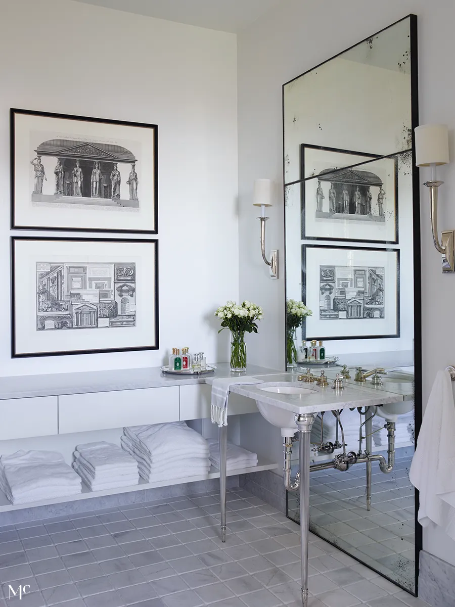 A bathroom with large black-and-white framed architectural prints on the wall, silver mirrors, a marble sink, a grey tile floor, and white linens