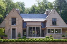 front exterior view of a traditional stone and tan brick home in a pristine, harmoniously designed style, with a blue roof, modern farmhouse aesthetic, large windows, and dark grey shutters, surrounded by lush green trees