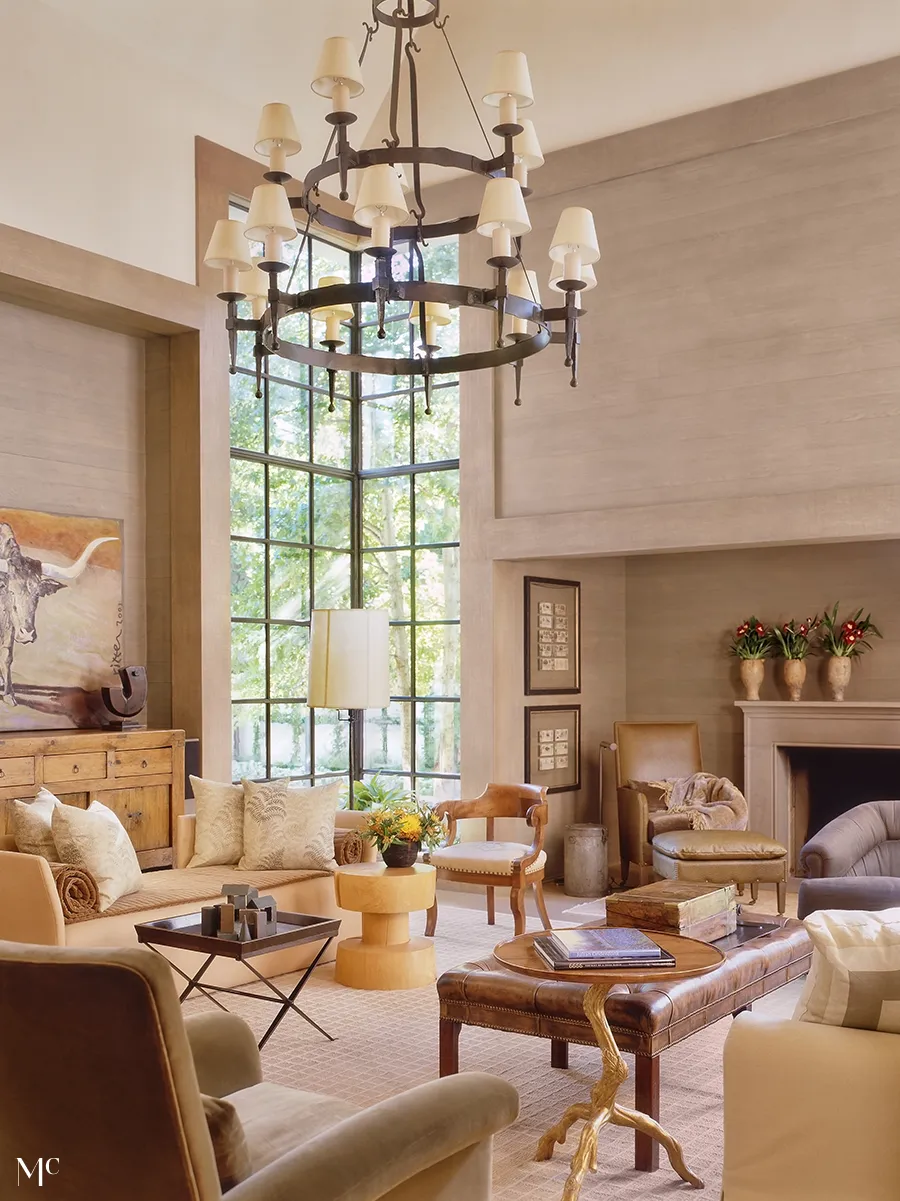 large windows with natural light, a grand chandelier above the sofa and armchairs in beige leather, wooden coffee table near fireplace, rustic textures, a wall painting of longhorn on one side, modern art pieces, warm lighting creating an inviting atmosphere