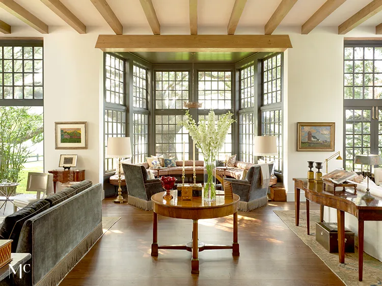 A large living room with tall windows, a sofa and armchairs in the center of an open space that leads to two dining areas, one on each side of the room, with wooden tables, a modern interior design style with rustic touches, and natural light from the large glass windows