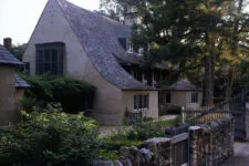 front and side view of an old, rustic cottage with a gray shingle roof surrounded by stone walls, an ivy-covered garden fence, and a black wooden gate leading to a courtyard