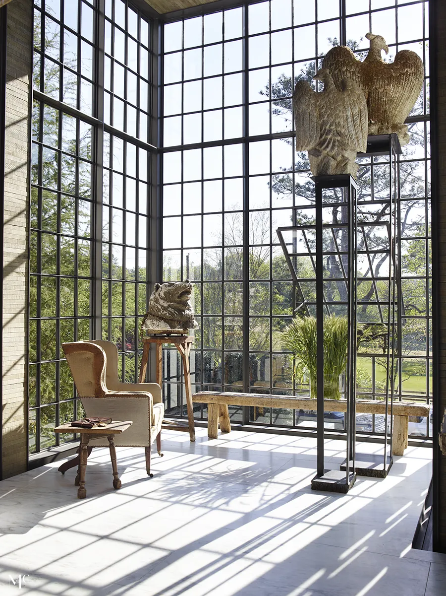 A modern living space with floor-to-ceiling black steel windows, wooden furniture, and sculptures that cast natural light and shadows, framed by a view of trees and nature outside.