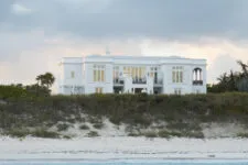 A wide shot of a white beach mansion in with simple design, sand dunes, and the ocean in the background