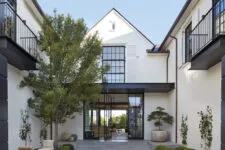 An architectural front view of a modern farmhouse courtyard with white and black buildings, large glass doors, oversized trees, and neutral tones