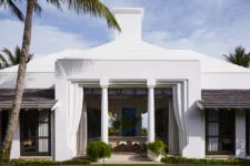 A front facade of an all-white bungalow with large windows, columns flanking each door, a shingled roof, circular chimney, and tropical plants on a green lawn