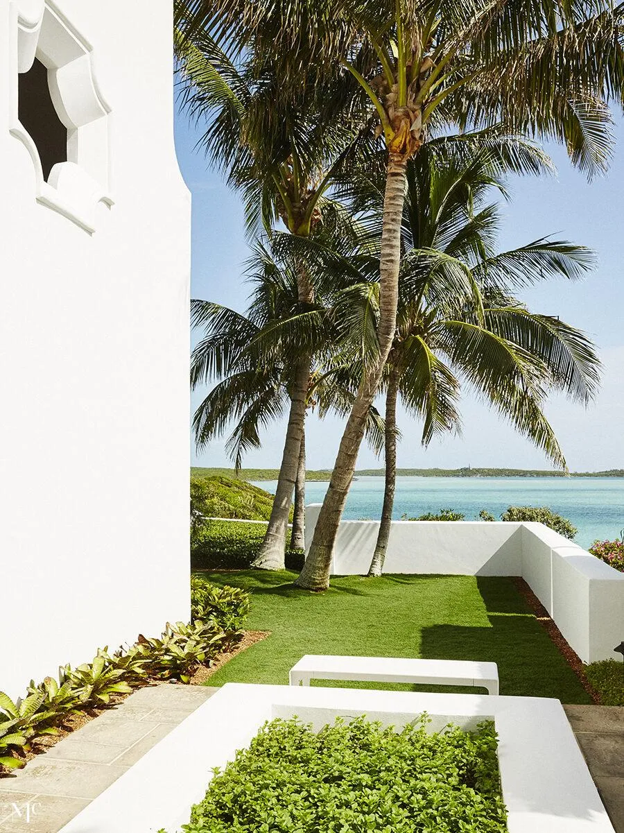 A front facade of an all-white bungalow with large windows, columns flanking each door, a shingled roof, circular chimney, and tropical plants on a green lawn