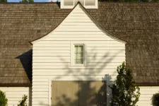 An architectural photograph of the exterior front view of an old white barn with a gable roof and a large garage door, surrounded by trees.