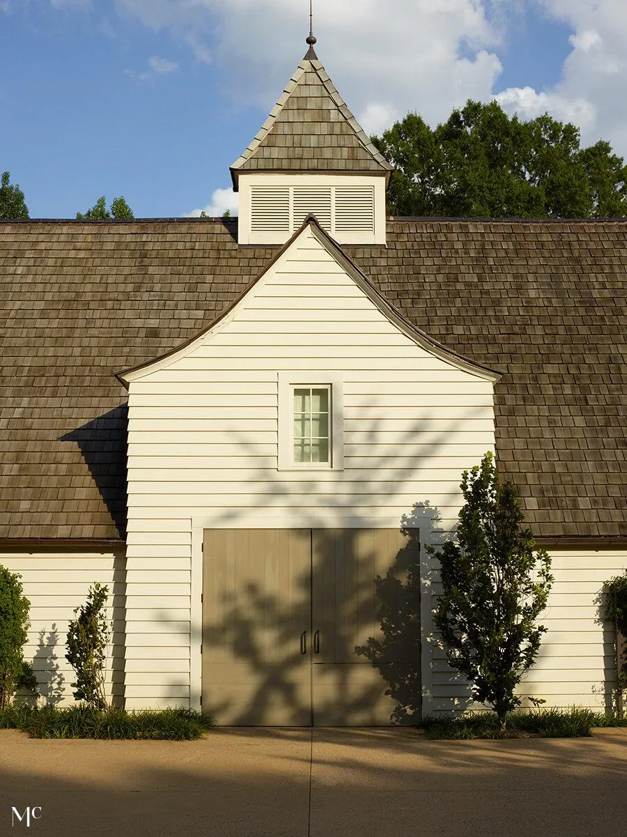 An architectural photograph of the exterior front view of an old white barn with a gable roof and a large garage door, surrounded by trees.